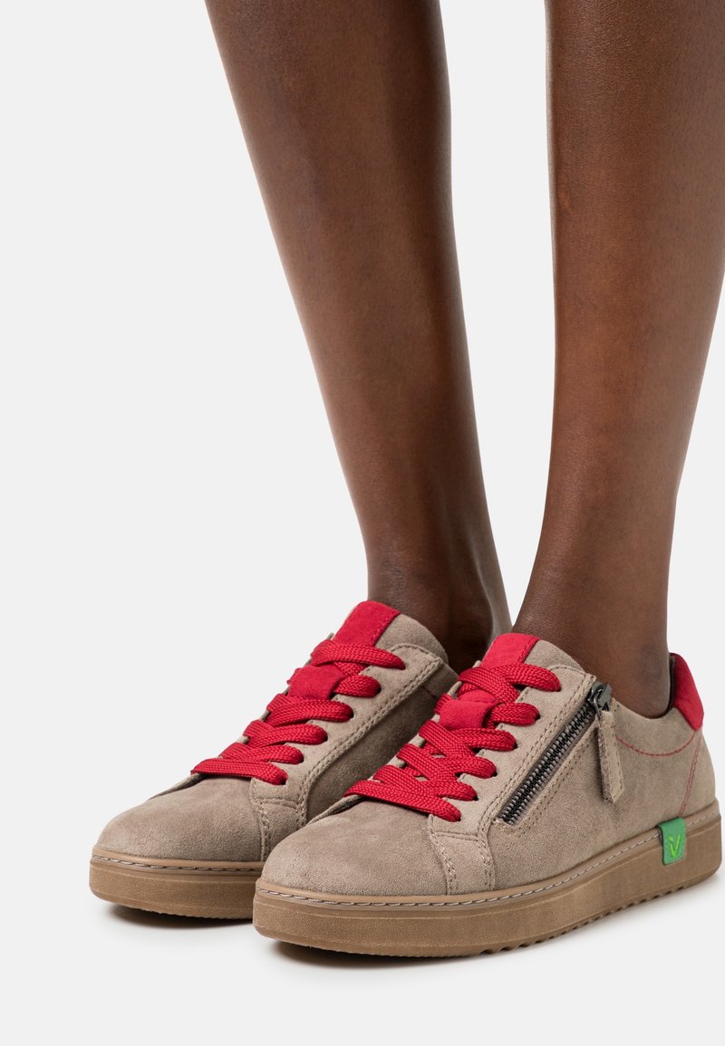 Sneaker in suede beige con lacci rossi, cerniera laterale e accento del logo verde. Presenta una spessa suola in gomma marrone e dettagli cuciti.