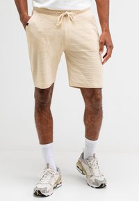 Beige Shorts aus strukturiertem Stoff, mit einem elastischen Bund und Kordelzug sowie Seitentaschen, getragen mit weißen Socken und Turnschuhen.