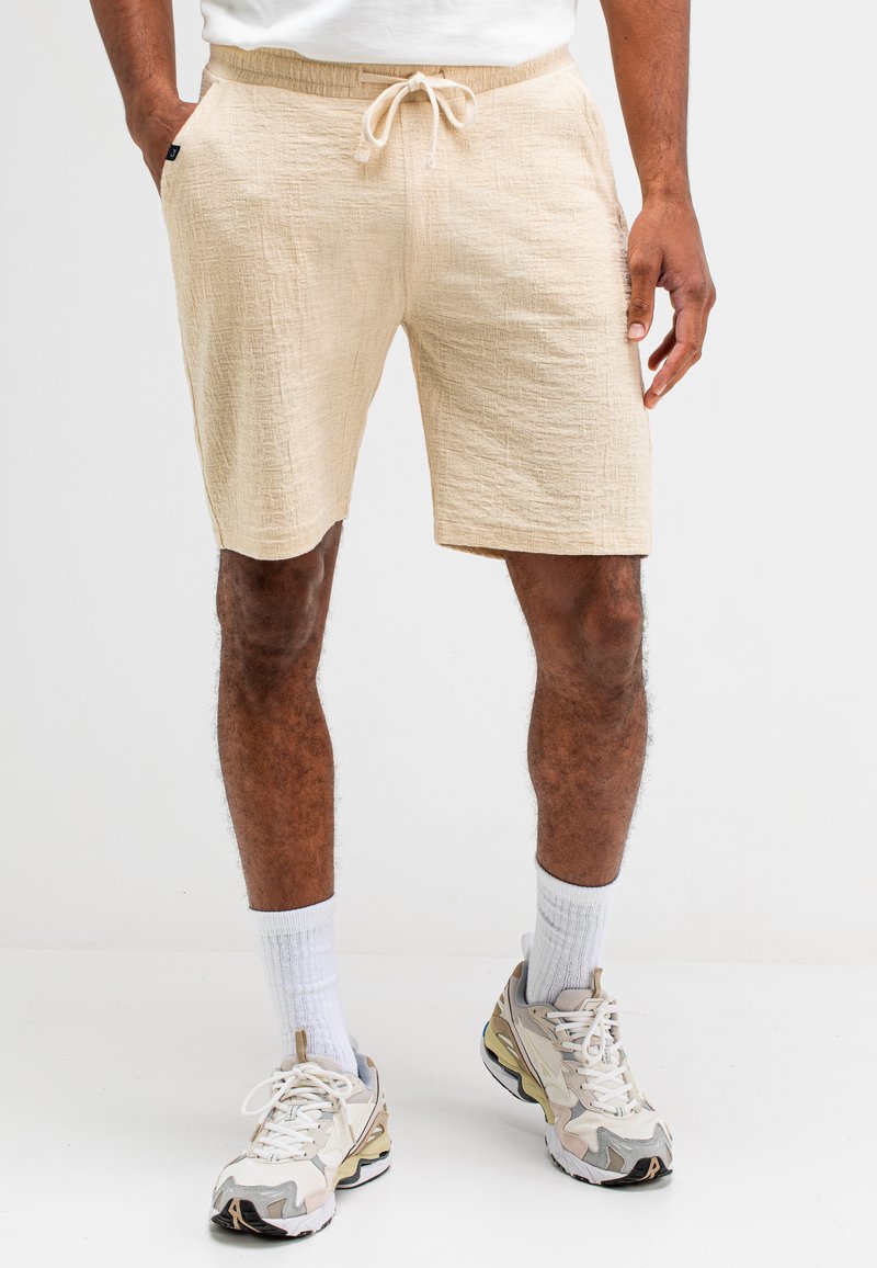 Beige Shorts aus strukturiertem Stoff, mit einem elastischen Bund und Kordelzug sowie Seitentaschen, getragen mit weißen Socken und Turnschuhen.