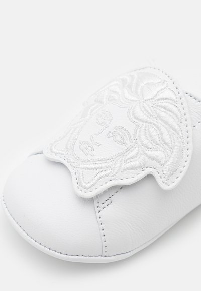 Hvitt skinnsko med et fremtredende brodert design med et klassisk ansiktsmotif. Glatt tekstur og synlige sømdetaljer.