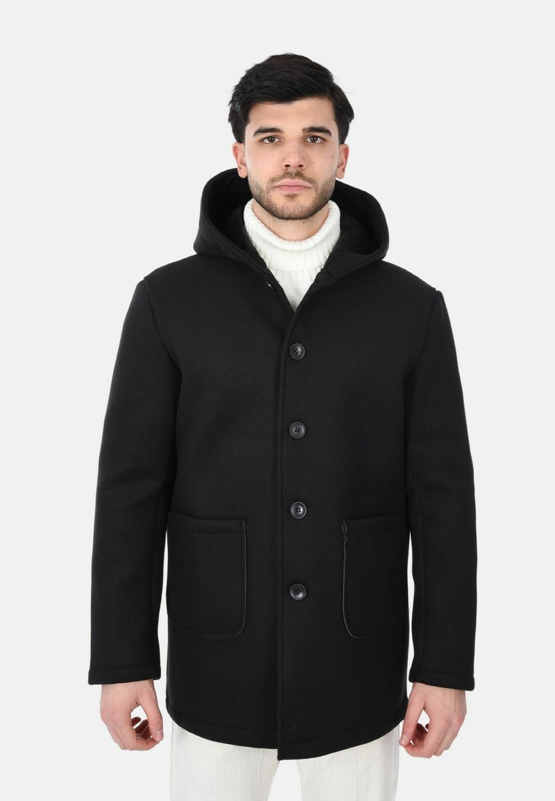 Cappotto nero con cappuccio in misto lana, con chiusura a bottoni, due tasche frontali e un maglione a collo alto a costine, testurizzato, indossato sotto.