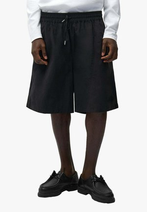 Uomo che indossa pantaloncini neri con coulisse fino al ginocchio, camicia bianca a maniche lunghe e scarpe nere con lacci, in piedi davanti a uno sfondo bianco.