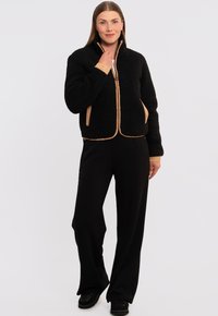 Giacca in pile nera con superficie testurizzata, rifiniture a contrasto in beige, chiusura con zip frontale e tasche laterali. Indossata con pantaloni neri a gamba larga.