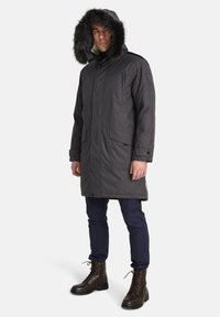 Parka grigia scura con cappuccio foderato in pelliccia sintetica, dotata di due tasche frontali e polsini regolabili, indossata sopra pantaloni blu scuro e stivali marroni con lacci.