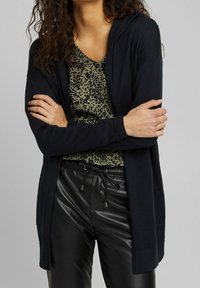 Black cardigan with a hood, long sleeves, and open front. Worn over a green animal print top and black leather trousers with drawstrings.
