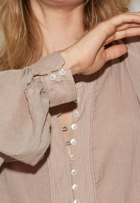 Blusa color marrone chiaro con maniche plissettate e texture, dotata di una chiusura frontale con bottoni perlacei e scollatura a V.