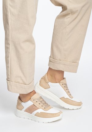 Person wearing beige corduroy pants and white sneakers with beige and light brown suede accents, standing on a white background.