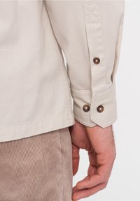 Camisa beige de manga larga con tela texturizada, con puños abotonados y detalle de costura reforzada en el brazo.