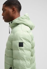 Giacca a vento verde chiaro con cappuccio, caratterizzata da un design trapuntato, una toppa nera sul braccio e chiusura con cordino sul collo.