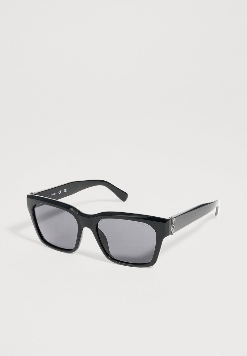 Lunettes de soleil rectangulaires noires avec verres teintés foncés et branches épaisses, reposant inclinées sur une surface unie gris clair.