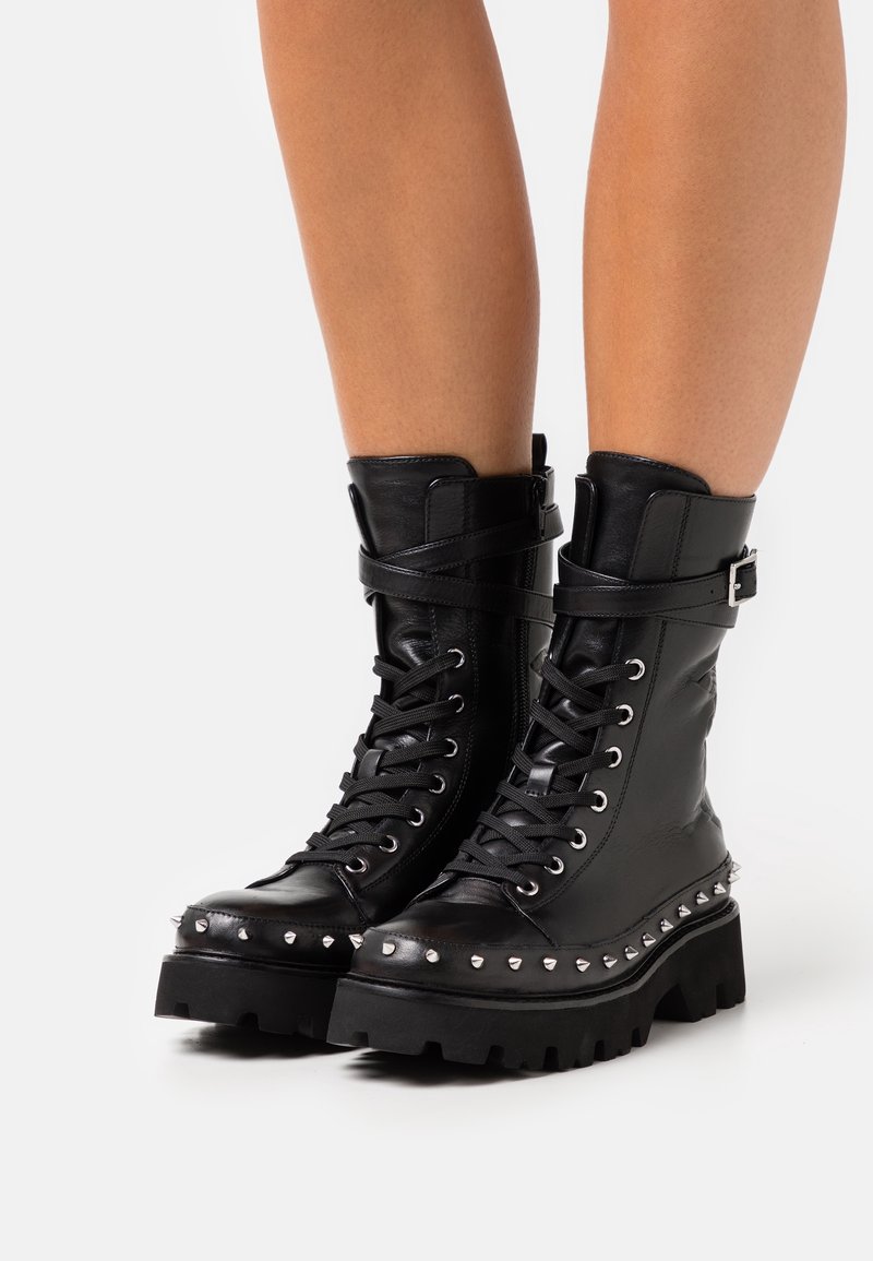Bottes de combat en cuir noir à lacets avec bordure cloutée et sangles à boucles portées sur des jambes nues, debout sur une surface blanche.