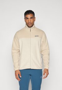 Beige fleecejakke med høj krave, fuld lynlås og logo på brystet. Blød tekstur og lange ærmer, med sidelommer og elastiske manchetter.