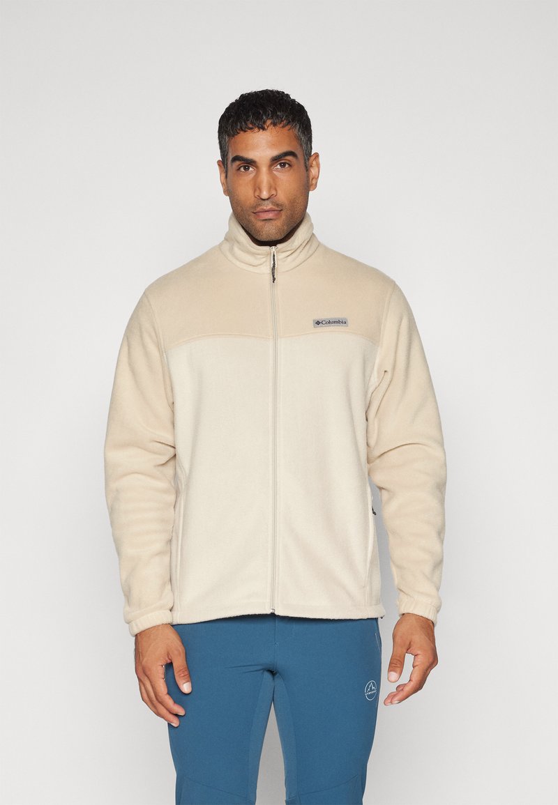 Beige fleecejakke med høj krave, fuld lynlås og logo på brystet. Blød tekstur og lange ærmer, med sidelommer og elastiske manchetter.