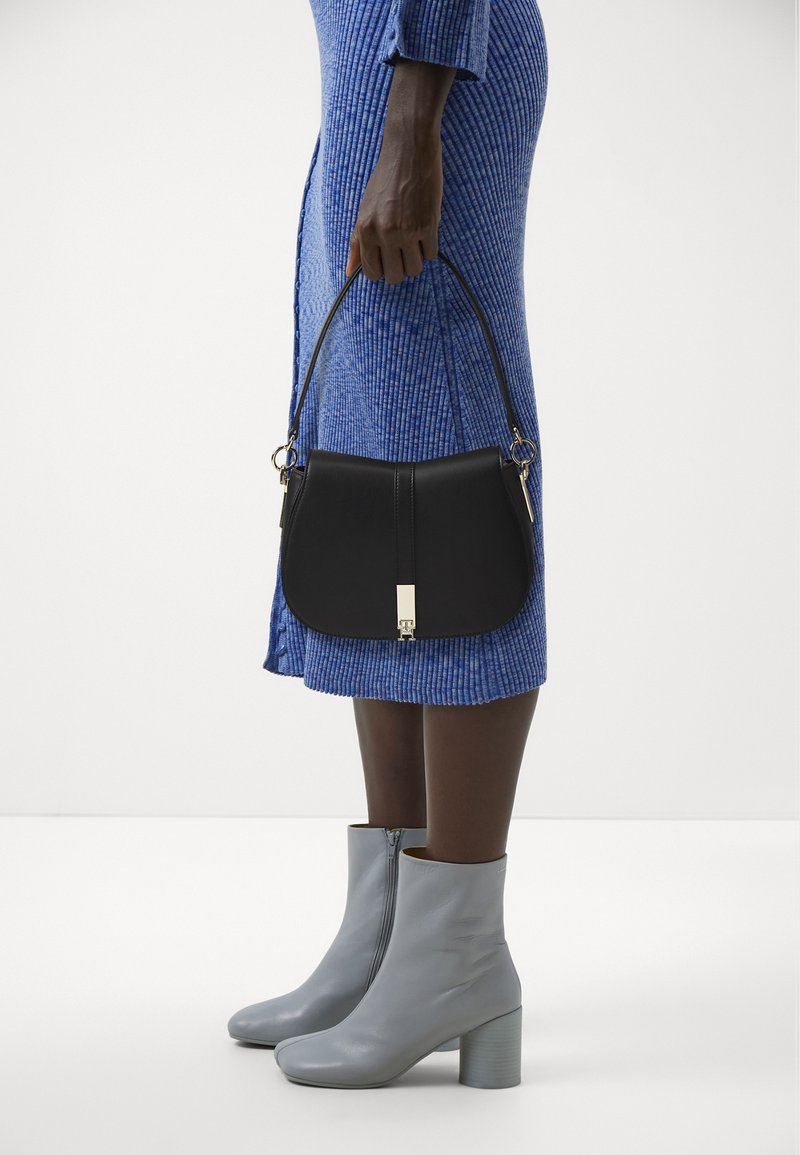 Personne en robe bleue côtelée tient un sac à main en cuir noir et porte des bottines gris clair à talons carrés sur un fond blanc.