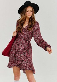 Krátke kvetinové zavinovacie šaty v tmavo ružovej farbe s bielymi a zelenými akcentmi, dlhé rukávy, volánový lem a zladená bordová taška.