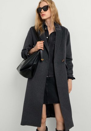 Femme portant un long manteau gris foncé, une jupe en denim noire, une chemise noire et des lunettes de soleil, tenant un sac à épaule en cuir noir.