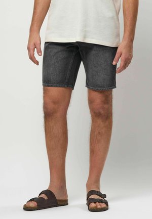 Muž v čiernych rifľových šortkách a čiernych sandáloch so sponou, stojaci s rukami uvoľnenými pozdĺž tela na svetlom jednofarebnom pozadí.