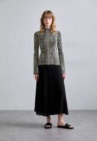 Black and white striped long-sleeve top paired with a black pleated maxi skirt and flat black sandals, standing on a gray floor.