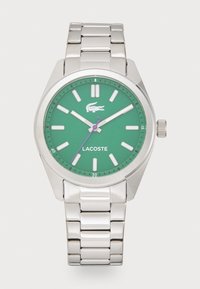 Ceas Lacoste cu un cadran verde, brățară din oțel inoxidabil argintiu, marcaje albe pentru ore și secundar violet. Include un logo și un indicator de dată.
