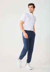 White short-sleeve button-up shirt, navy drawstring joggers, and white sneakers. Smooth fabric, relaxed fit, with elastic cuffs on pants.