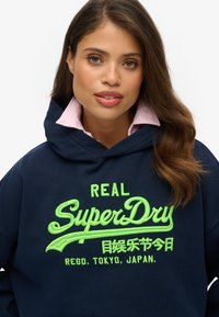Sudadera azul marino con el logotipo bordado en verde brillante de "SuperDry", con una textura suave y una camisa de color rosa con cuello visible debajo.