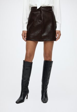 Femme portant une minijupe en cuir marron foncé et des bottes en cuir noires à talons montant jusqu'aux genoux sur fond blanc.