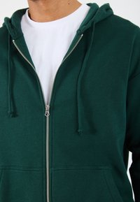 Veste à capuche zippée verte avec une poche avant, un cordon de serrage sur la capuche et une fermeture éclair argentée. Portée par-dessus un t-shirt blanc uni. Matière en coton avec une texture lisse.