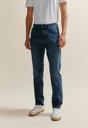 Jean en denim bleu foncé avec une coupe droite, cinq poches et une taille mi-haute, associés à des baskets blanches avec une semelle en caoutchouc marron.