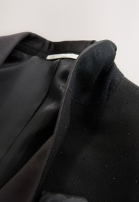Cuello de chaqueta negra hecha a medida con pequeñas cuentas decorativas y forro interior suave, mostrado plegado abierto sobre un fondo claro.