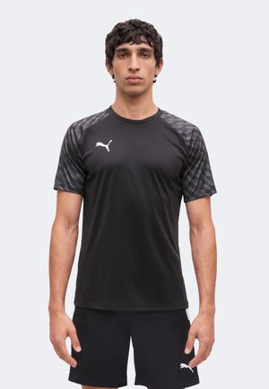 Bărbat stă în picioare, cu fața înainte, purtând un tricou sport negru Puma cu mâneci cu model și pantaloni scurți negri Puma asortați, pe un fundal simplu și deschis la culoare.