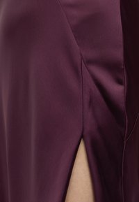 Tissu satin lisse violet foncé avec une fente latérale révélant la peau claire en dessous, mettant en valeur la texture du vêtement et les détails des coutures.