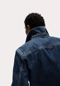 Chaqueta de denim con un acabado azul oscuro, que presenta un collar alto, costuras visibles y un corte relajado. El fondo es claro y neutro.