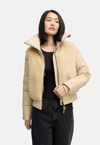Beige gewatteerde jas met een pluizige textuur kraag, ritssluiting aan de voorkant, geribbelde manchetten en een cropped ontwerp. G gedragen over een zwarte top.
