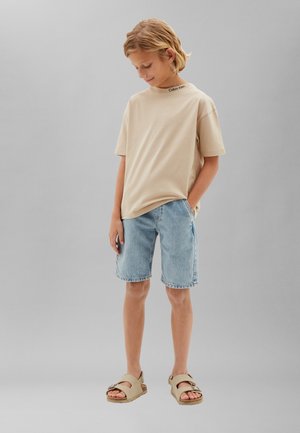Dieťa so svetlými vlasmi, ktoré nosí béžové tričko Calvin Klein, svetlomodré rifľové šortky a béžové sandále, stojí s jednou rukou v vrecku.