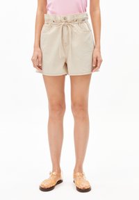 Mujer vistiendo pantalones cortos de cintura tipo paperbag en beige, atados con un cordón, una blusa rosa y sandalias de gel transparentes, de pie contra un fondo blanco.