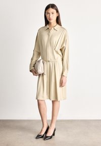 Žena v béžových šatách s dlhými rukávmi a opaskom držiaca zladenú tašku, stojaca na leštenej betónovej podlahe, obutá v čiernych lodičkách.