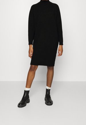 Person iført en sort langærmet turtleneck-kjole med sorte ankelstøvler og hvide sokker, stående på en ensfarvet grå gulv.
