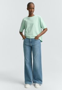 T-shirt oversize verde chiaro con maniche corte, abbinata a jeans blu a vita alta e a gamba larga. Il modello si trova in piedi con le mani nelle tasche.