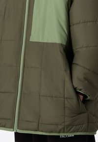 Olívazöld és világoszöld pufi kabát. Vízszintes steppelt mintával és kontrasztos részletekkel rendelkezik. Masszív cipzárral és zsebekkel ellátva.