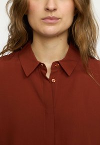 Rust-colored button-up blouse with a collar, featuring orange buttons, a smooth texture, and neat stitching.