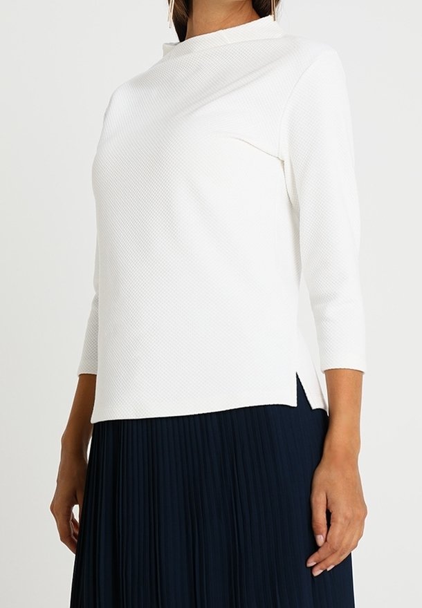 Femme portant un haut blanc texturé à manches longues avec un col montant et une fente latérale, associé à une jupe plissée bleu marine.