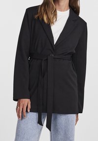 Černé sako s páskem v pase, dlouhými rukávy, klopovým límcem, jednobotonovým zapínáním a hladkým materiálem, spárované se světlými džíny.