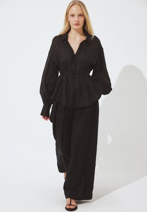 Blondínka kráča, oblečená v voľnej čiernej blúzke so zviazaným pásom, širokých čiernych nohaviciach a čiernych sandáloch s remienkami na bielom pozadí.