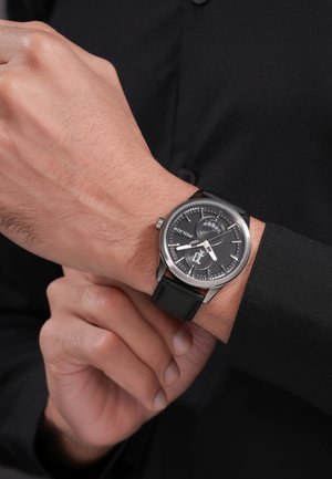 Laikrodis su juodu odiniu dirželiu ir juodu tekstūruotu ciferblatu, rodantis laiką, nešiojamas ant kairės riešo, dešinė ranka reguliuoja dirželį, matomas juodas marškinių rankovė.