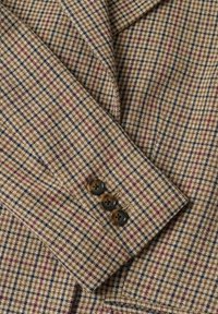 Tissu marron à carreaux avec un motif de petits carrés en tons crème, bordeaux et bleu. Présente trois boutons foncés sur le poignet.