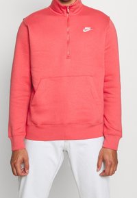 Nike zip-up-tröja i ljus korall, med hög krage, långa ärmar och en framlänges känguruficka. Mjuk tygstruktur.