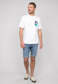 T-shirt en coton blanc avec un graphique coloré de palmier sur la poche, associé à un short en denim retroussé et des chaussures slip-on beige clair.