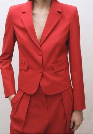 Vrouw draagt een nauwsluitend rood colbert met twee knopen en bijpassende hooggetailleerde rode broek met plooien, staand tegen een effen achtergrond.