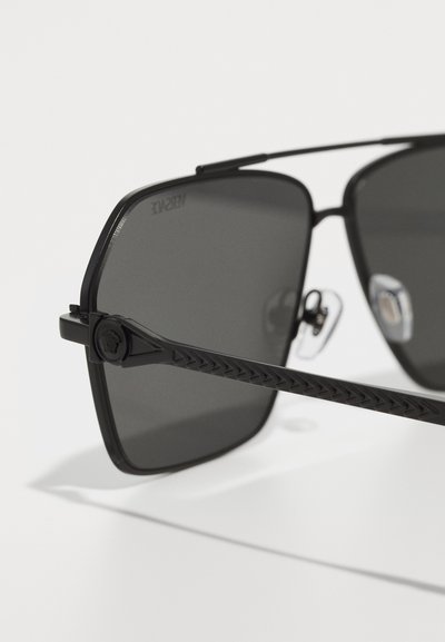 Lunettes de soleil noires avec des verres de forme carrée, verres teintés foncés, monture en métal noir élégant, branches noires texturées et plaquettes de nez ajustables.