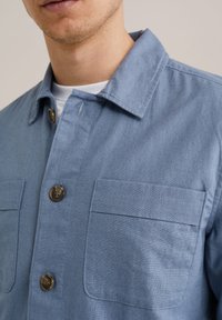 Camisa de botón azul claro hecha de suave tela de mezclilla, con un cuello clásico, dos bolsillos en el pecho y botones marrones. Textura suave.